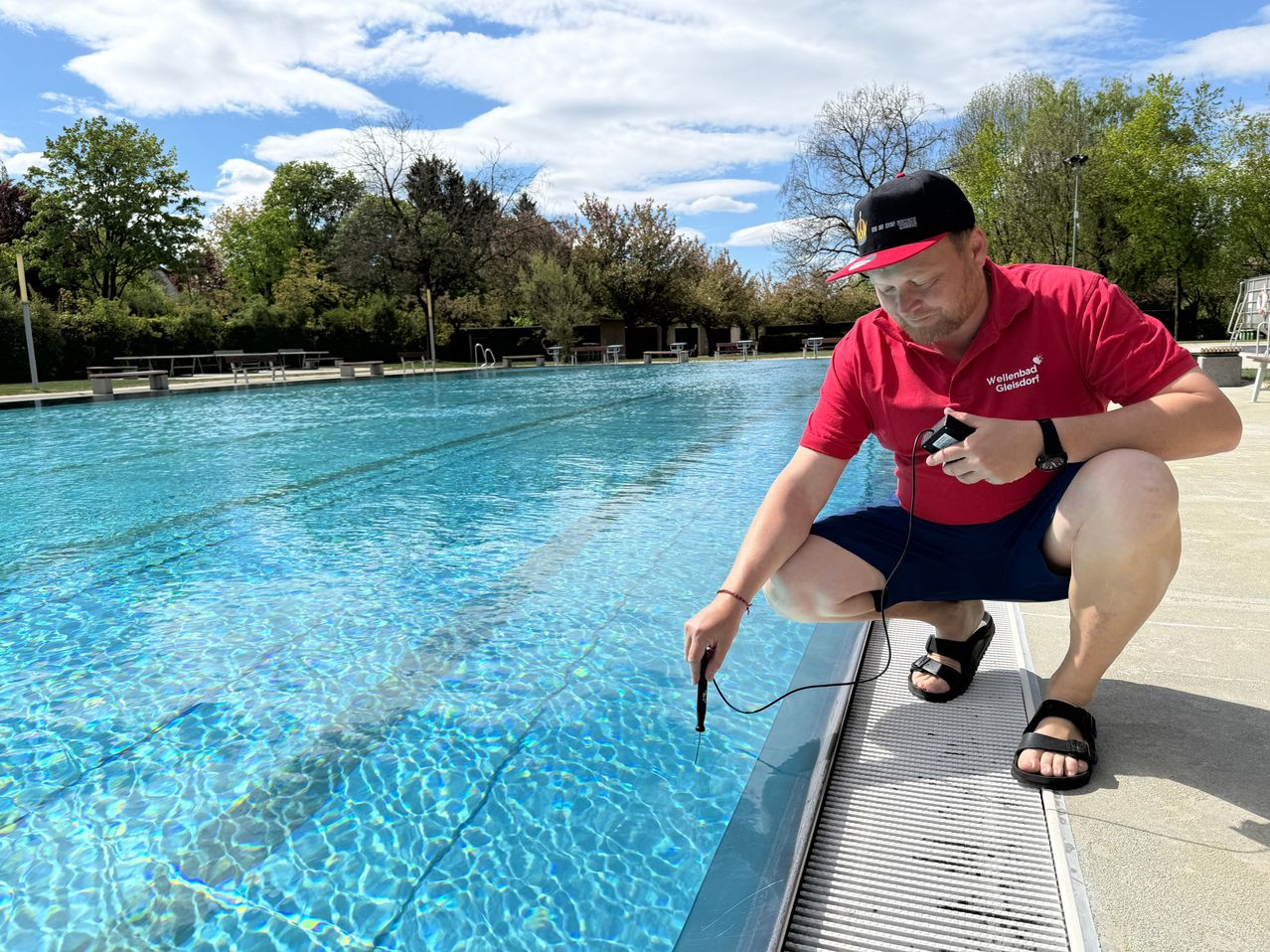 Ein Mann überprüft die Wasserqualität in einem Freibad, trägt ein rotes Shirt und einen schwarzen Hut.