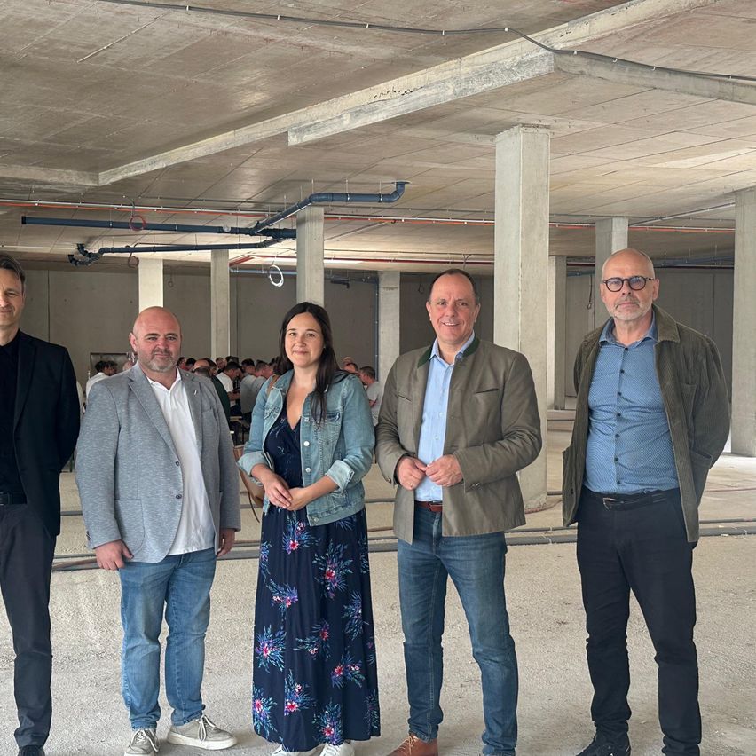 Five people are standing in an unfinished building, perhaps for a photo. There are large pillars, pipes, and cables on the ceiling. In the background, there are several people sitting on chairs.