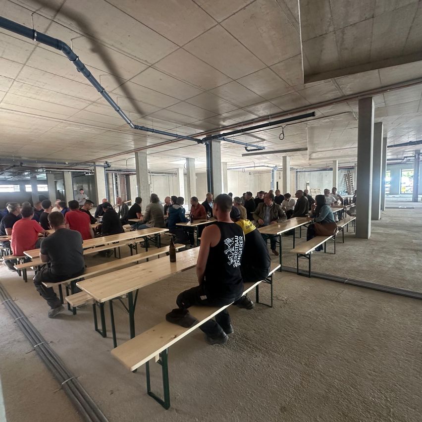 A large indoor area with several people sitting on benches, possibly a factory canteen. The room has metal pipes on the ceiling.