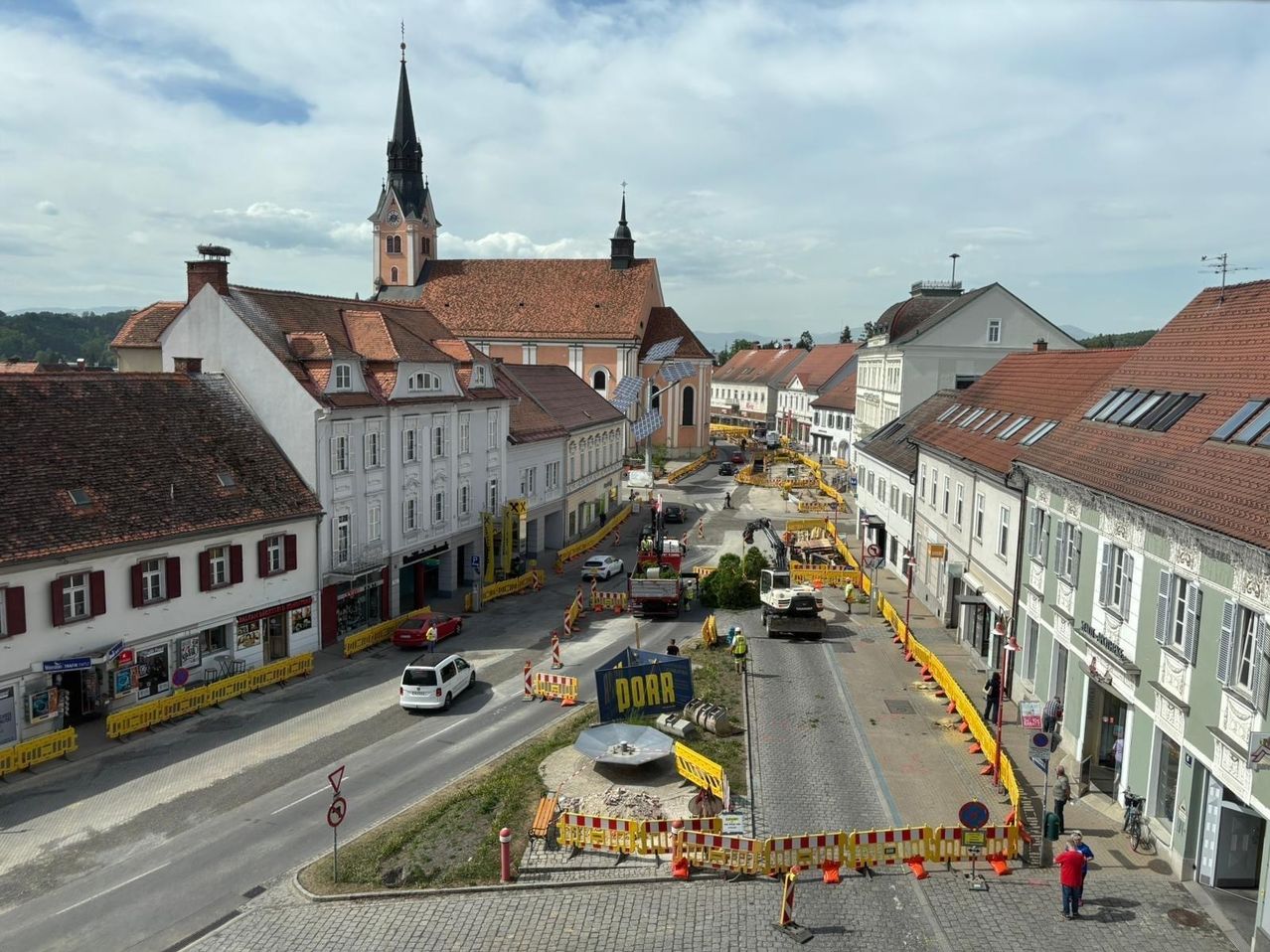 Aerial view of a town with ongoing road construction. Buildings, cars, and construction barriers visible. A church tower is in the background.