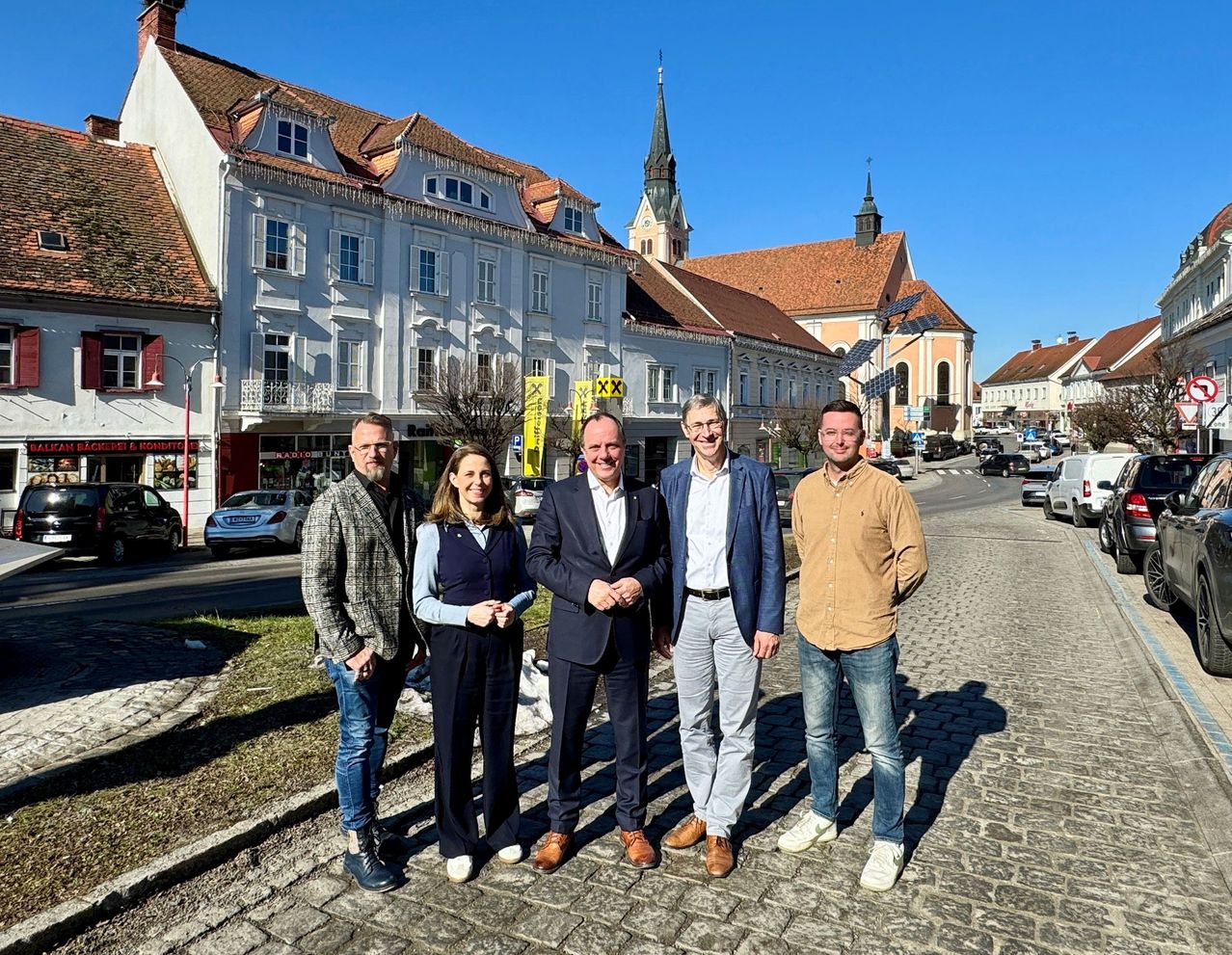 Fünf Personen stehen auf einer Kopfsteinpflasterstraße und lächeln für ein Foto. Hinter ihnen befinden sich Gebäude, Autos und eine Kirche mit Solarpanelen.