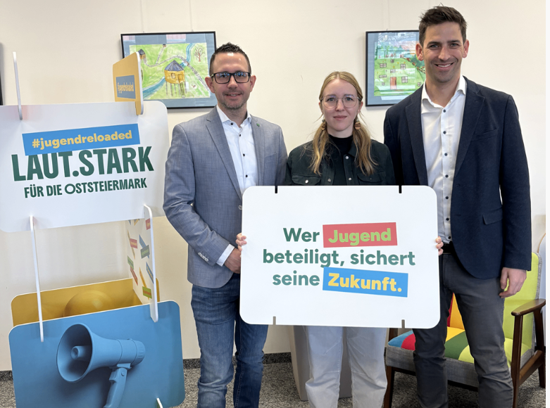 Drei Personen stehen vor einem Banner, zwei Männer und eine Frau, lächelnd. Sie halten ein Schild mit der Aufschrift 'Wer Jugend beteiligt, sichert seine Zukunft'. Hinter ihnen steht ein farbenfroher Stuhl, und auf der linken Seite befindet sich ein Megaphon. An der Wand hängen zudem gerahmte Bilder.