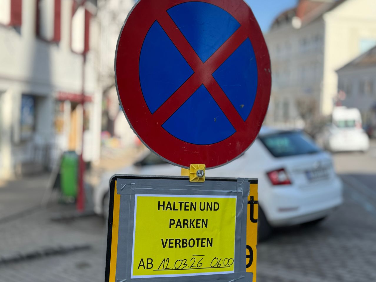 Ein Schild verbietet das Halten und Parken ab 12.03.26 um 06:00. Ein weißer Wagen ist geparkt.