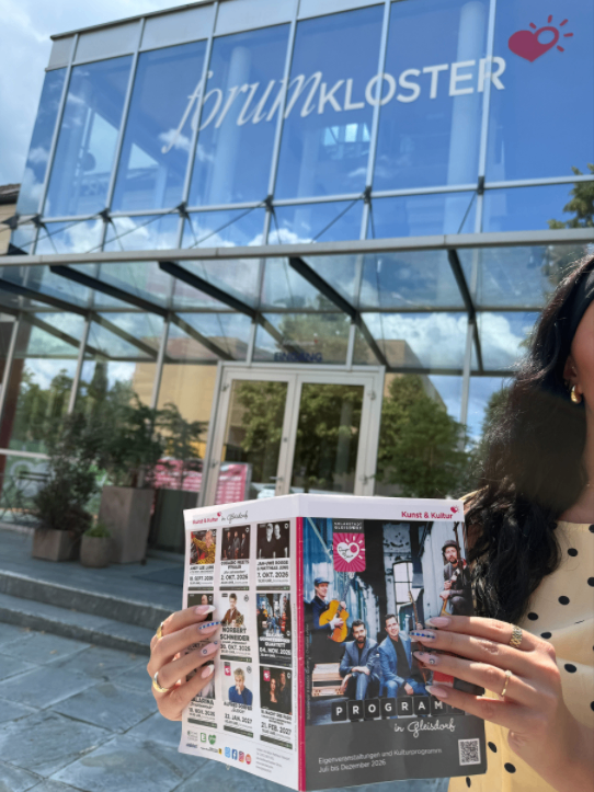 Eine Frau hält eine Zeitschrift mit einer bunten Titelseite, auf der Musiker abgebildet sind und der Text 'PROGRAMM in Gleisdorf' steht, vor einem modernen Gebäude mit einer Glaswand und einem Herzlogo.