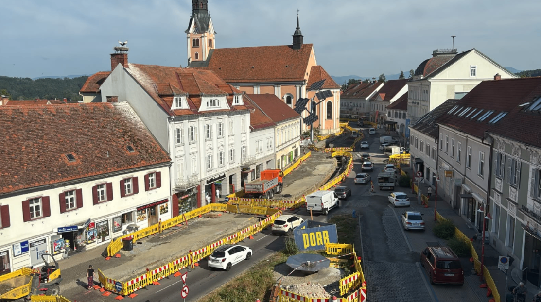 Luftaufnahme einer Stadt mit laufenden Straßenbauarbeiten, mit mehreren Fahrzeugen, Gebäuden und einer Kirche im Hintergrund.