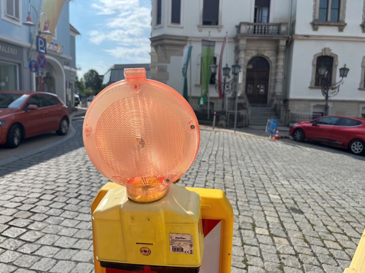 Ein gelber und oranger Verkehrskegel mit einer reflektierenden orangefarbenen Spitze steht auf einer Kopfsteinpflasterstraße. Im Hintergrund ist ein weißes Gebäude mit Balkon und Fahnen zu sehen.