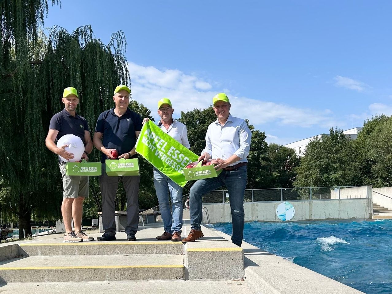 Vier Männer in passenden gelben Hüten stehen auf Stufen neben einem Pool, halten Boxen und eine grüne Fahne mit der Aufschrift 'Apfel essen nicht vergessen'. Sie posieren für ein Foto.