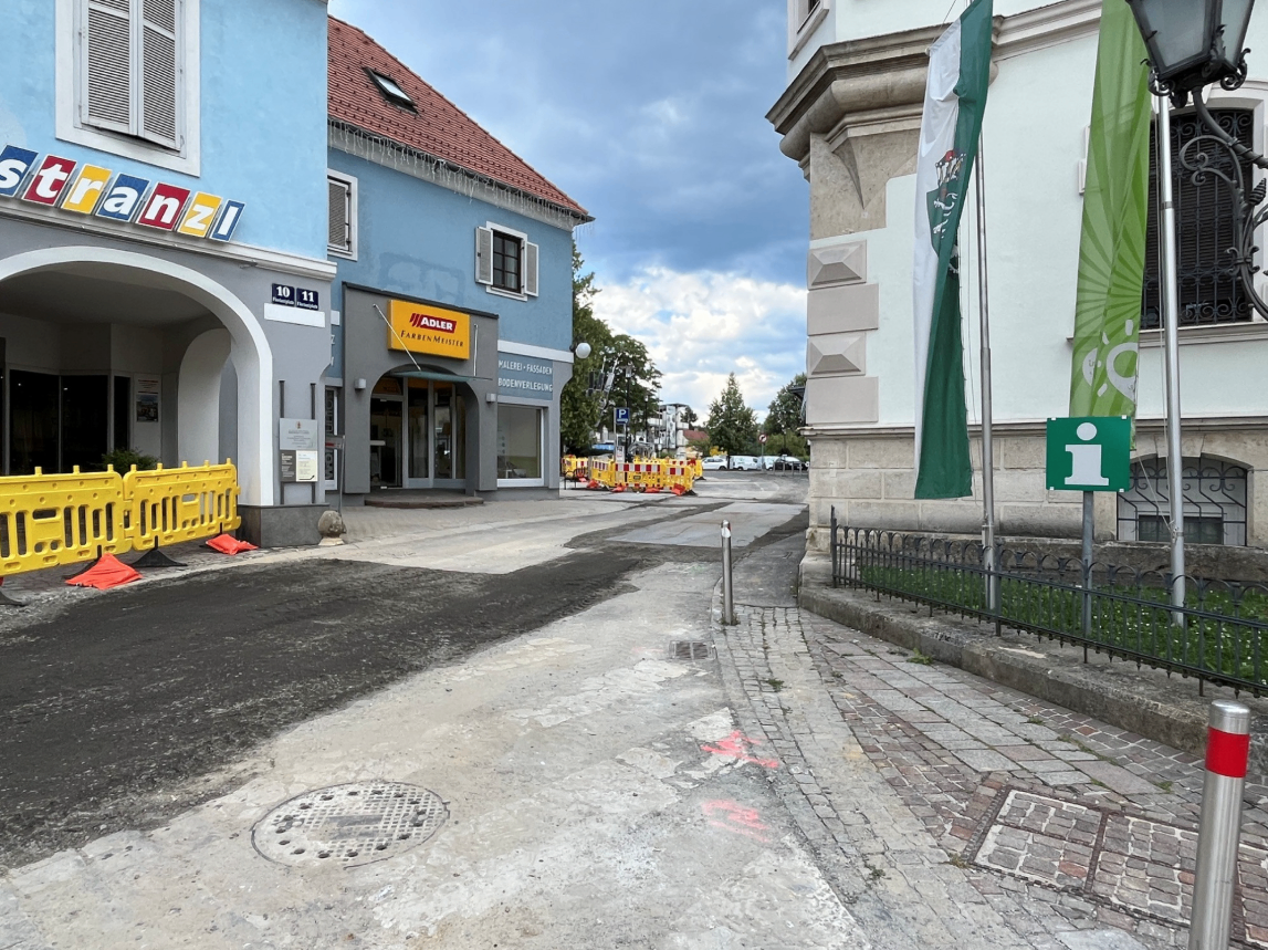 Eine Straße mit einer Baustelle vor einem blauen Gebäude mit einem gelben Schild, das Adler sagt. Es gibt Absperrungen und ein grünes Schild mit dem Buchstaben i.