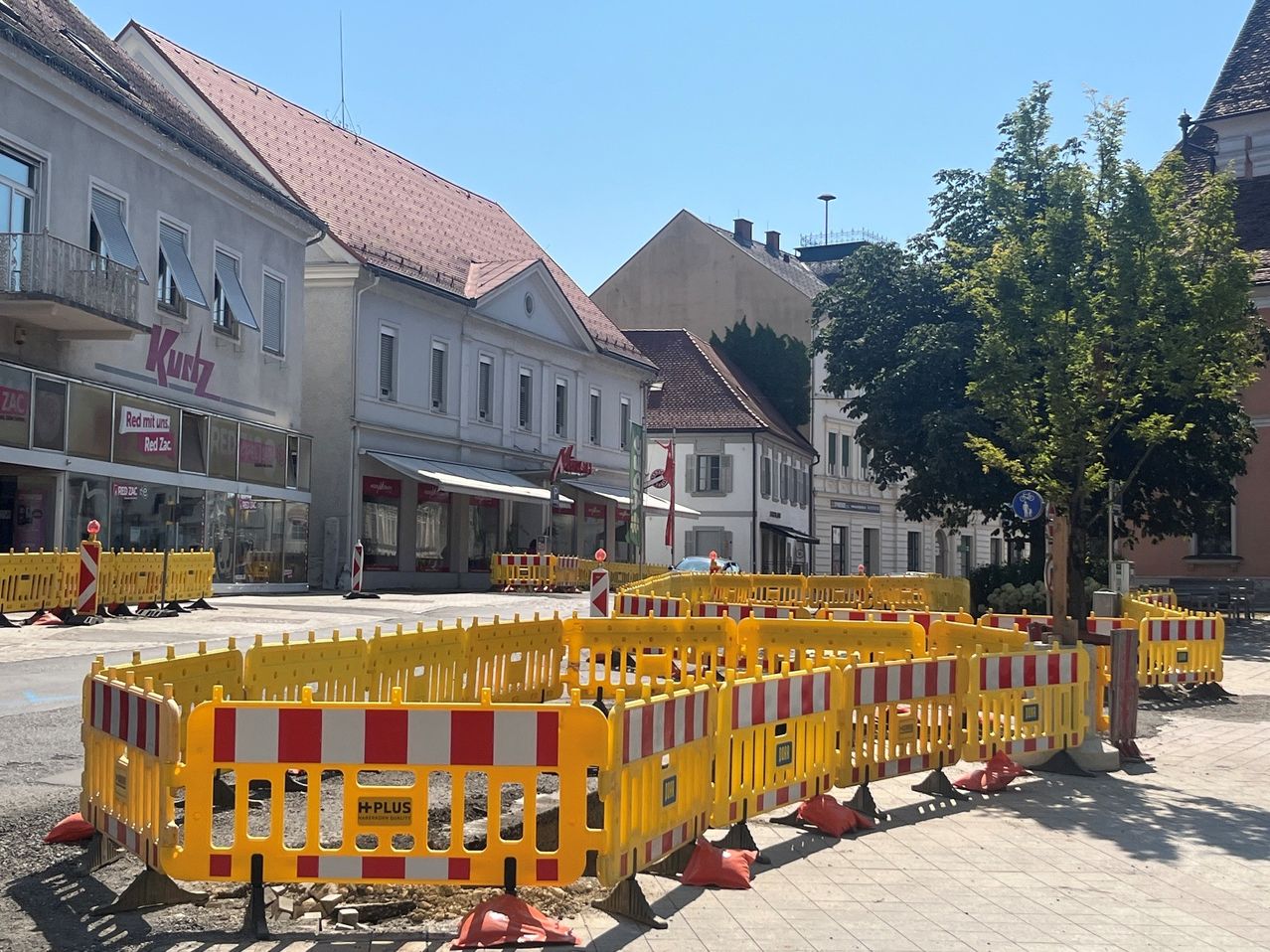 Eine Stadtstraße ist mit gelben und roten Barrikaden abgesperrt. Im Hintergrund sind Gebäude mit Geschäften und ein Baum zu sehen.