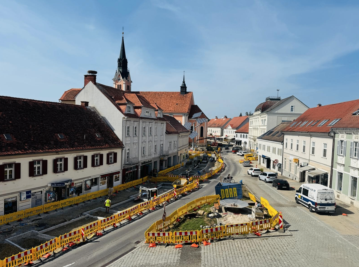 Luftaufnahme einer Stadt mit laufenden Straßenbauarbeiten. Gelbe Barrieren und Maschinen sind sichtbar. Gebäude mit roten Dächern und einem Turm im Hintergrund. Autos und ein Lieferwagen fahren auf der Straße.