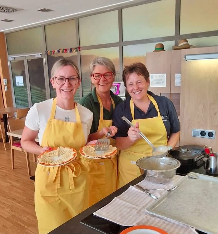 Drei Frauen in gelben Schürzen stehen in einer Küche, lächeln und halten Platten mit Waffeln. Eine Frau hält eine Pfanne, und eine andere hält einen Kochlöffel. Die Küche hat einen Herd, ein Waschbecken und einen Tisch mit einer Schüssel.