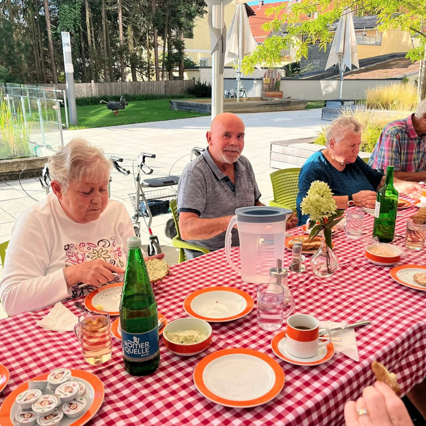 Eine Gruppe älterer Menschen sitzt um einen Tisch mit rot-weiß kariertem Tischtuch und genießt eine Mahlzeit. Auf dem Tisch stehen Teller, Tassen und Flaschen. Im Hintergrund befindet sich ein Gebäude, ein Rasenbereich und Bäume.