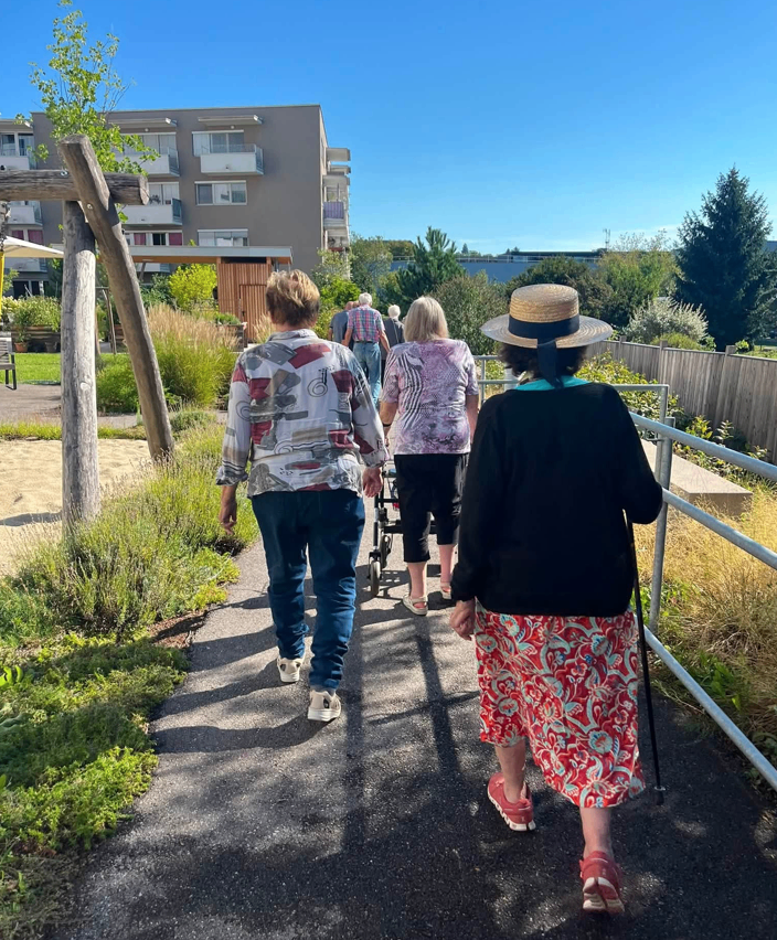 Eine Gruppe älterer Frauen geht auf einem gepflasterten Weg in einem Park. Hinter ihnen befindet sich ein Gebäude mit Balkonen.