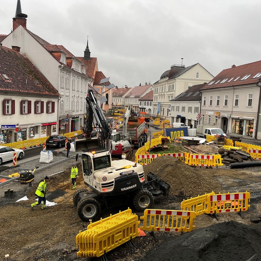 Eine Baustelle mit Maschinen und Arbeitern in einer Stadt. Ein großer Bagger gräbt die Straße aus. In der Nähe gibt es Erdhaufen und Bauzäune.