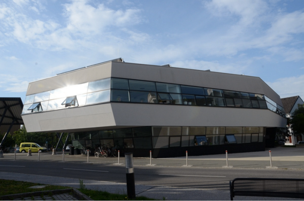 Ein modernes Gebäude mit großen Glasfenstern, umgeben von geparkten Fahrrädern und Pollern vor dem Eingang.