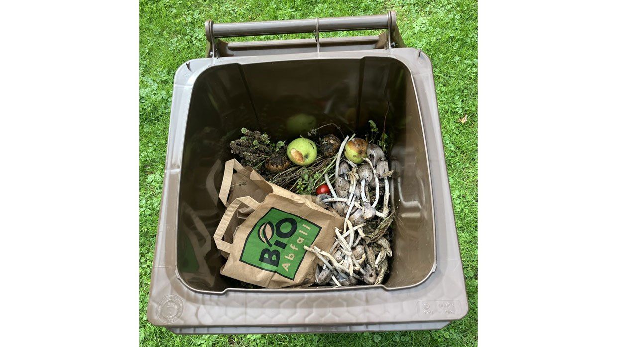 Ein Kompostbehälter gefüllt mit organischen Abfällen, einschließlich Fruchtschalen, Gemüseresten und Gartenabfällen, auf einer Grasfläche platziert.