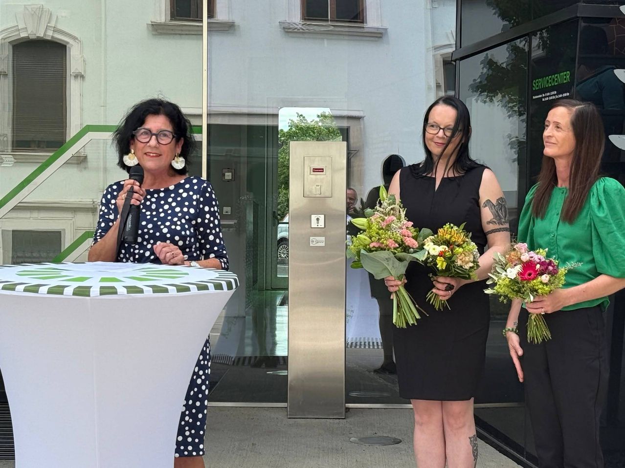 Drei Frauen, eine mit einem Mikrofon, stehen vor einem Gebäude. Sie halten Blumen und scheinen an einer Veranstaltung teilzunehmen. Dahinter zeigt eine Reflexion eine Glastür mit einer Pflanze.