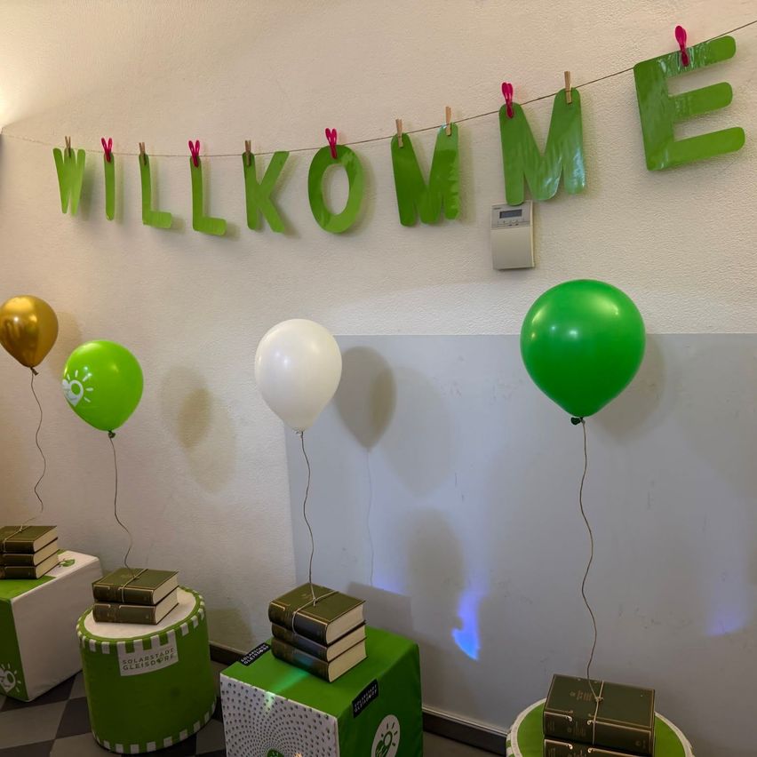 Ein Raum mit einer weißen Wand und einem grünen Banner mit der Aufschrift 'Willkommen'. Drei Ballons hängen über grünen Kisten mit Büchern darauf.