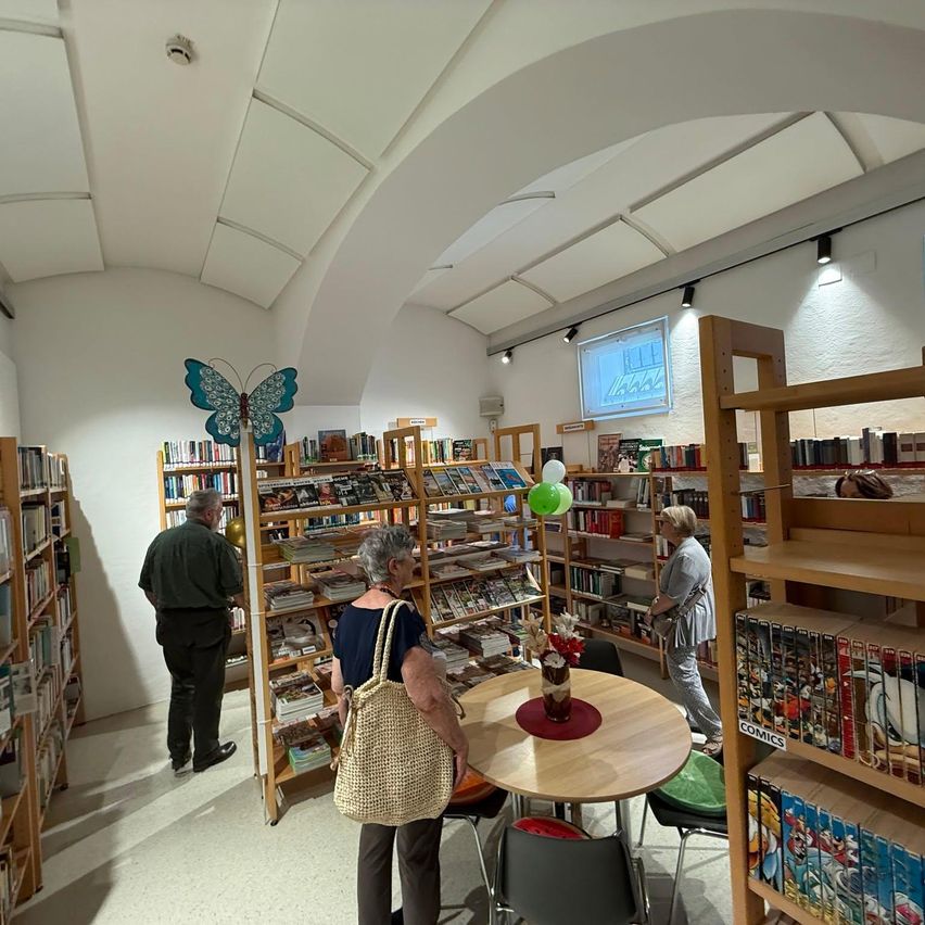 Eine Innenbuchhandlung mit mehreren Regalen voller Bücher. Zwei Frauen stöbern in Büchern, und ein Mann steht neben einem Bücherregal. In der Mitte steht ein Tisch mit einer Blumenvase.