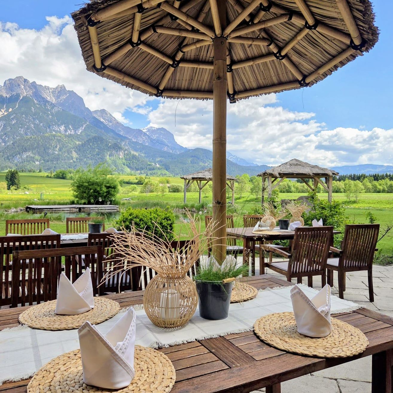 Ein Tisch mit zwei Gedecken und einem Bambus-Umbrella, in einem Restaurant mit Bergblick. Der Tisch hat dekorative Pflanzen und Servietten. Andere Tische und Stühle sind aufgestellt.