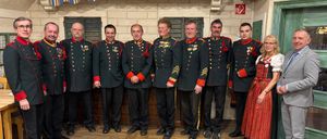 Neun Männer und eine Frau in traditionellen Uniformen stehen für ein Foto in einer Reihe. Dahinter befindet sich eine Wand mit einer Ziegelsteinstruktur und einem hölzernen Vordach.