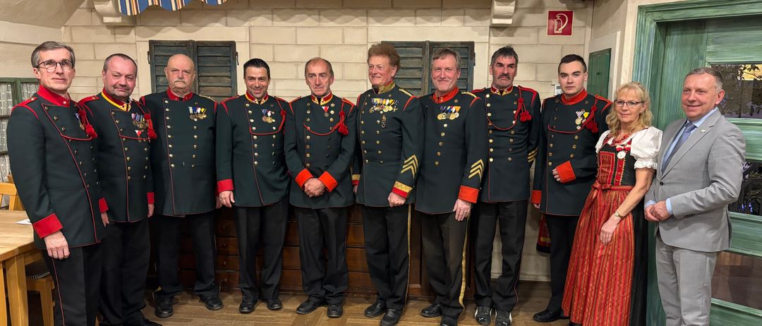 Neun Männer und eine Frau in traditionellen Uniformen stehen für ein Foto in einer Reihe. Dahinter befindet sich eine Wand mit einer Ziegelsteinstruktur und einem hölzernen Vordach.