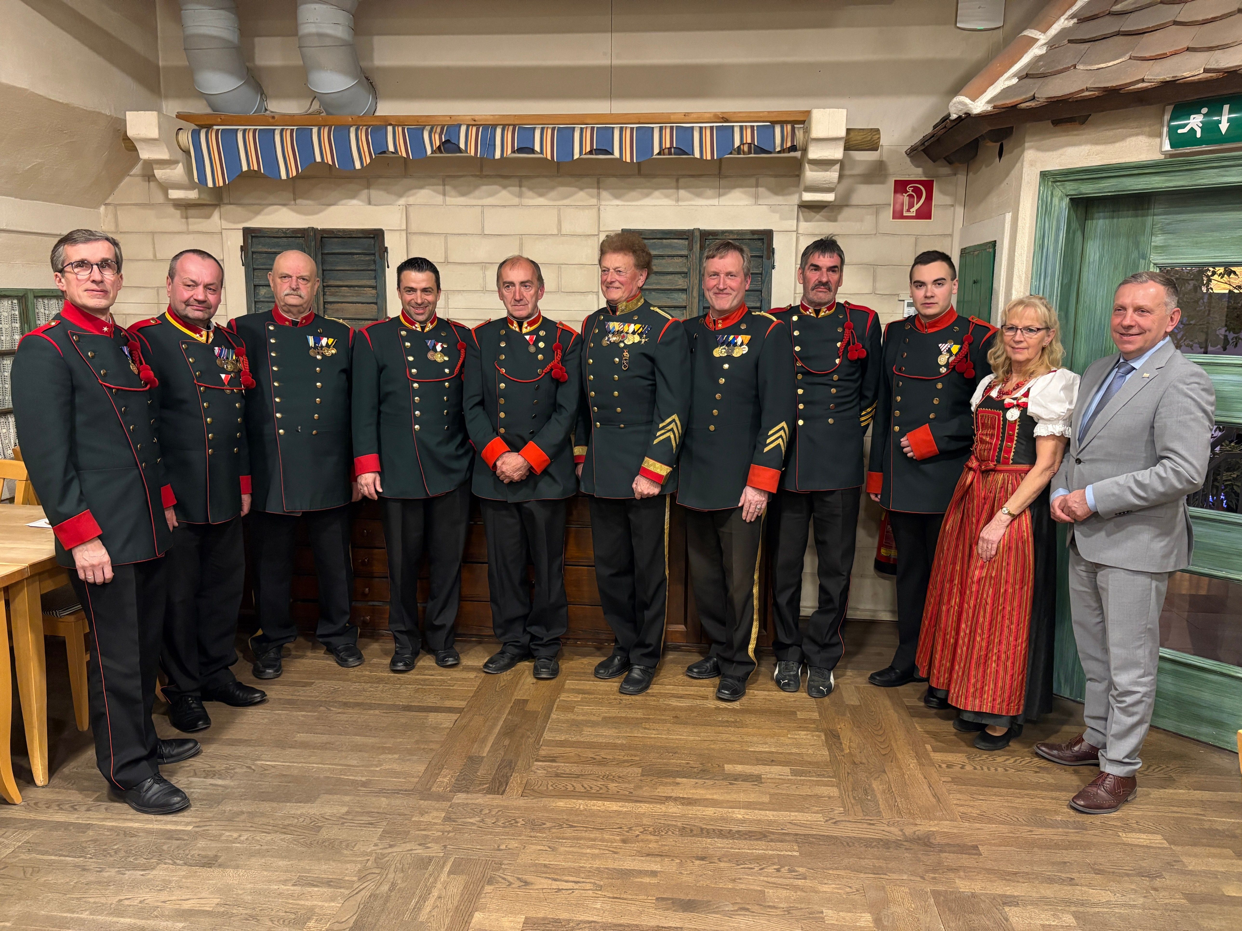 Eine Gruppe von Männern und einer Frau stehen in einer Reihe in Militäruniformen. Sie stehen vor einer mit Dekorationen geschmückten Wand. Sie alle lächeln und scheinen für ein Foto zu posieren.