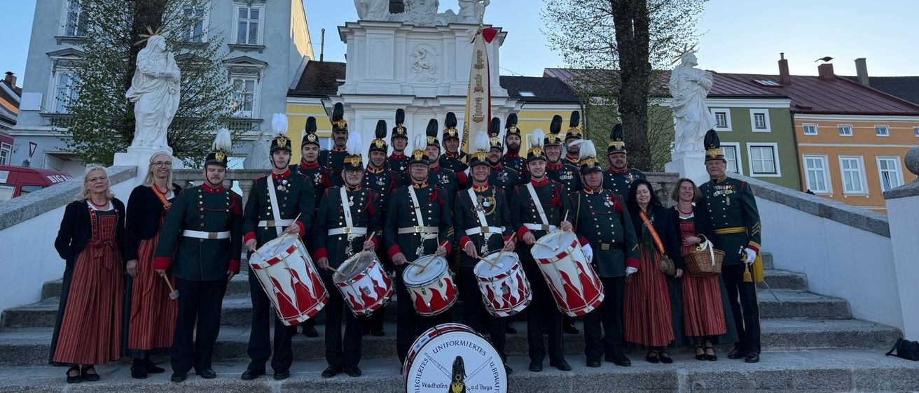 Ein Militärorchester posiert für ein Foto auf einem Stadtplatz, mit einer Statue hinter ihnen, einem roten Trommel in der Front, und Bäumen und Gebäuden rundherum.