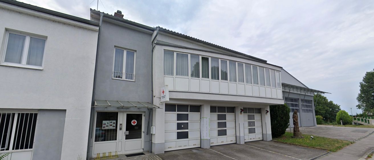 Ein zweistöckiges Gebäude mit mehreren Fenstern und drei Garagentoren. Der Eingang befindet sich auf der linken Seite und über ihm befindet sich ein rotes Kreuz-Schild.