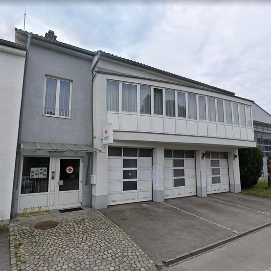 Die Vorderseite eines Gebäudes mit einem Schild, das ein rotes Kreuz zeigt. Es gibt drei Garagentore und einen kleinen Garten. Der Himmel ist bewölkt.