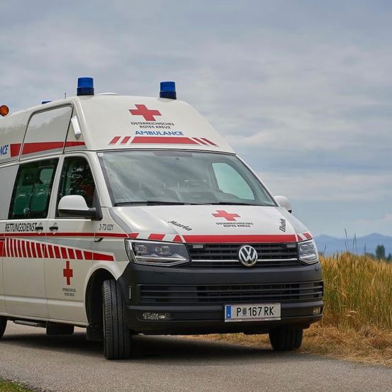 Ein weißer und roter Krankenwagen mit blauen Lichtern auf dem Dach fährt auf einer Straße. Das Fahrzeug hat das österreichische Rote-Kreuz-Emblem an der Seite.