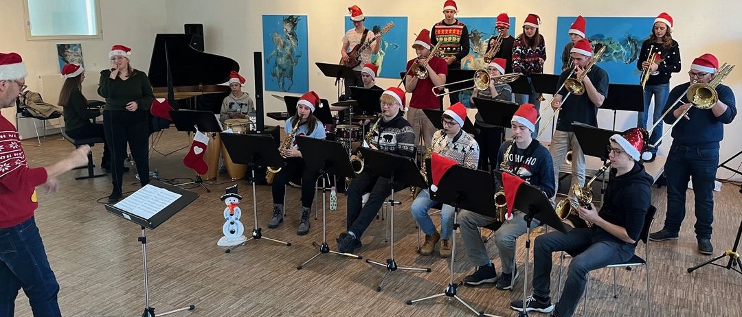Eine Gruppe Musiker in Weihnachtsmützen spielt mit verschiedenen Instrumenten wie Saxophonen, Trompeten und Trommeln in einem Raum mit einem Schneemann und einem Klavier.