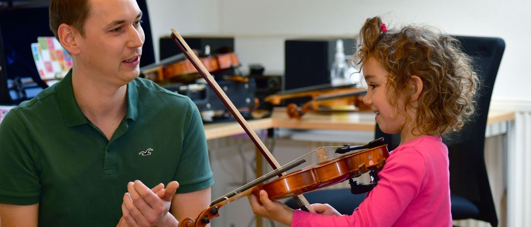Ein junger Junge in einem grünen Poloshirt spielt die Geige mit einem kleinen Mädchen in einem rosa Kleid. Beide konzentrieren sich auf das Instrument. Dahinter sind Geigen ordentlich auf einem Schreibtisch angeordnet.