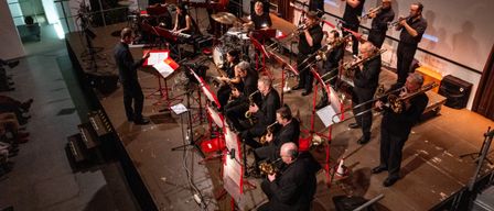 Eine Jazzband spielt auf einer Bühne vor einer Projektionsleinwand. Die Musiker sind in Schwarz gekleidet und spielen verschiedene Instrumente wie Trompeten, Saxophone und Schlagzeug. Einige stehen, andere sitzen, mit Notenständern vor ihnen.