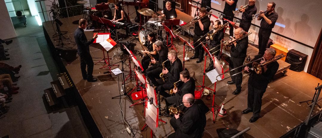 Eine Jazzband spielt auf einer Bühne vor einer Projektionsleinwand. Die Musiker sind in Schwarz gekleidet und spielen verschiedene Instrumente wie Trompeten, Saxophone und Schlagzeug. Einige stehen, andere sitzen, mit Notenständern vor ihnen.