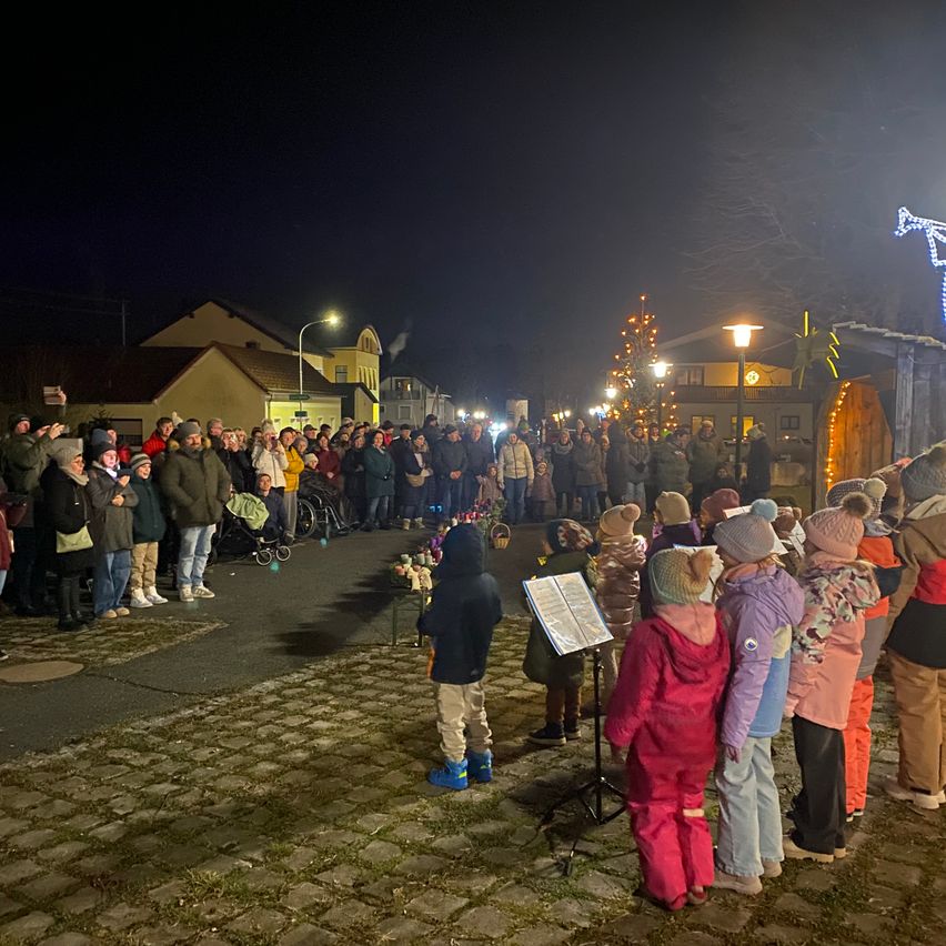 In der Nacht versammelt sich eine große Menschenmenge auf einer Kopfsteinpflasterstraße für eine Veranstaltung. Kinder stehen vorne mit einem Notenständer, und Erwachsene dahinter. Gebäude mit Weihnachtsdekoration säumen die Straße.