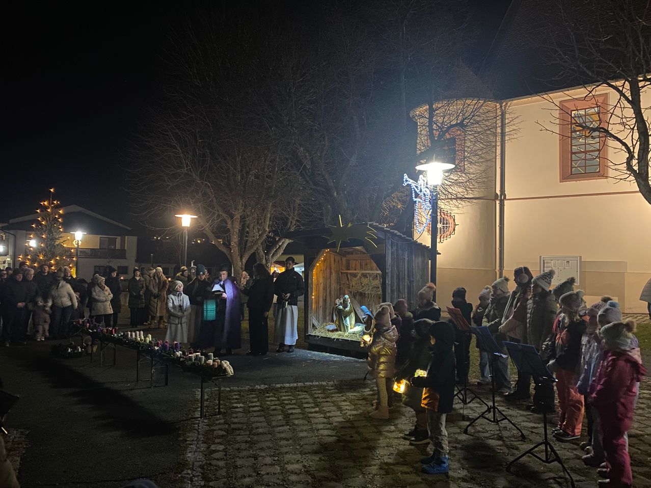 Eine Gruppe von Menschen versammelt sich bei Nacht um eine Krippe, hält Kerzen und singt. Die Szene wird von einer Straßenlaterne beleuchtet und von Bäumen umgeben.