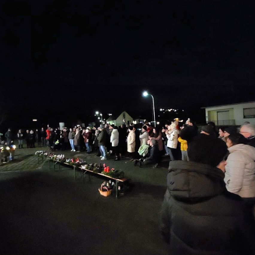 Viele Menschen haben sich bei Nacht versammelt, einige stehend, andere sitzend. Sie tragen Winterkleidung und halten Kerzen. Ein Tisch mit Blumen und Kerzen steht bereit. Straßenlaternen beleuchten die Szene.