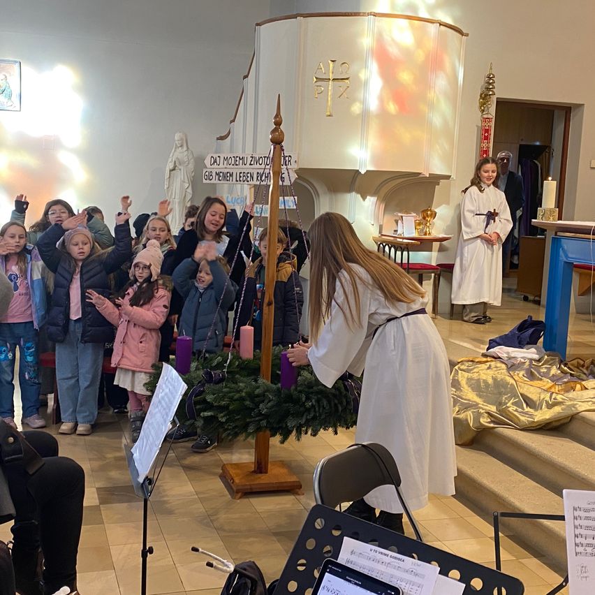 Eine Gruppe von Kindern steht um einen geschmückten Pfahl, singt und hält Kerzen. Eine Frau in einem weißen Gewand hält einen Kranz. Ein Schild mit deutschem Text ist an der Wand montiert. Eine Statue einer Frau und ein Kreuz befinden sich an der Wand.