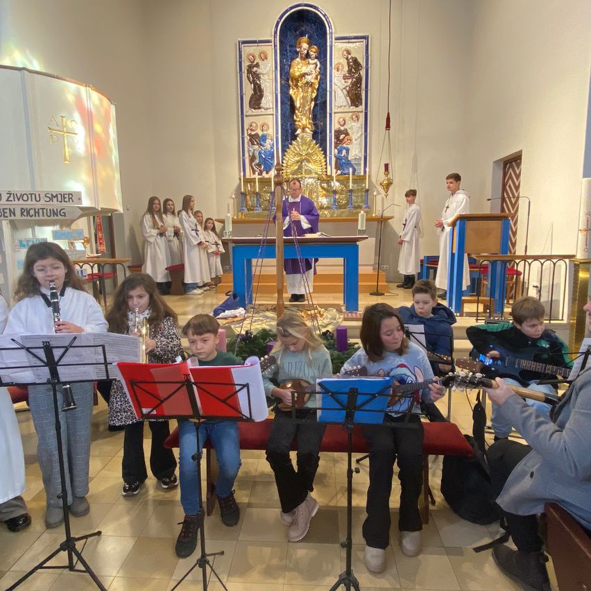 Eine Gruppe von Kindern spielt Instrumente in einer Kirche, mit zwei Priestern vor dem Altar. Ein Banner mit der Aufschrift 'Zivotu Smjer Ben Richtung' ist zu sehen. Die Kinder spielen Gitarren, Klarinetten und andere Instrumente.