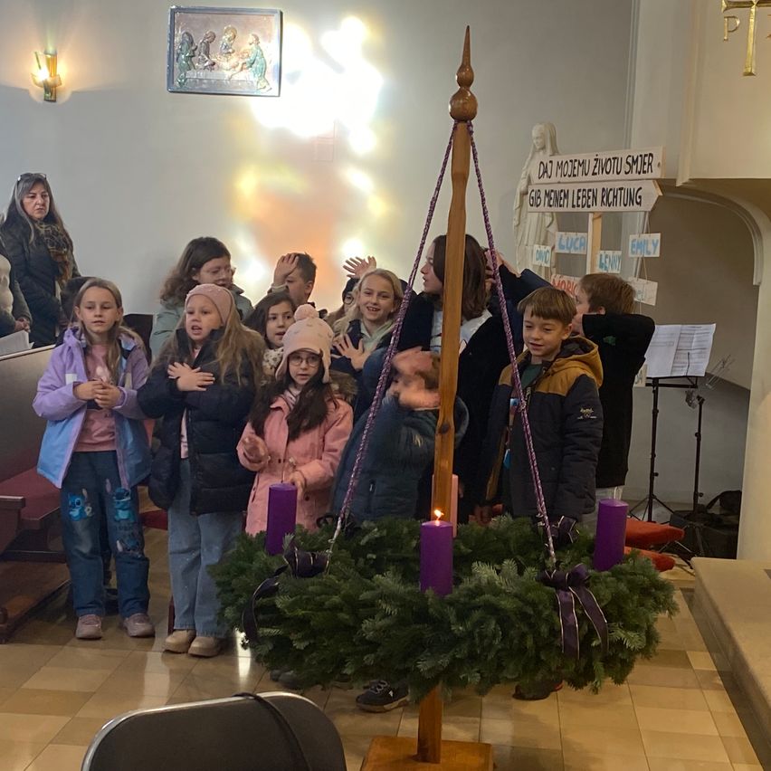 Kinder versammeln sich um einen kerzenbeleuchteten Adventskranz in einer Kirche, einige mit den Händen über den Herzen, andere halten Kerzen. Ein Holzpfahl hält den Kranz hoch. Ein Schild auf Deutsch lautet: 'Gib dem Leben Richtung.'