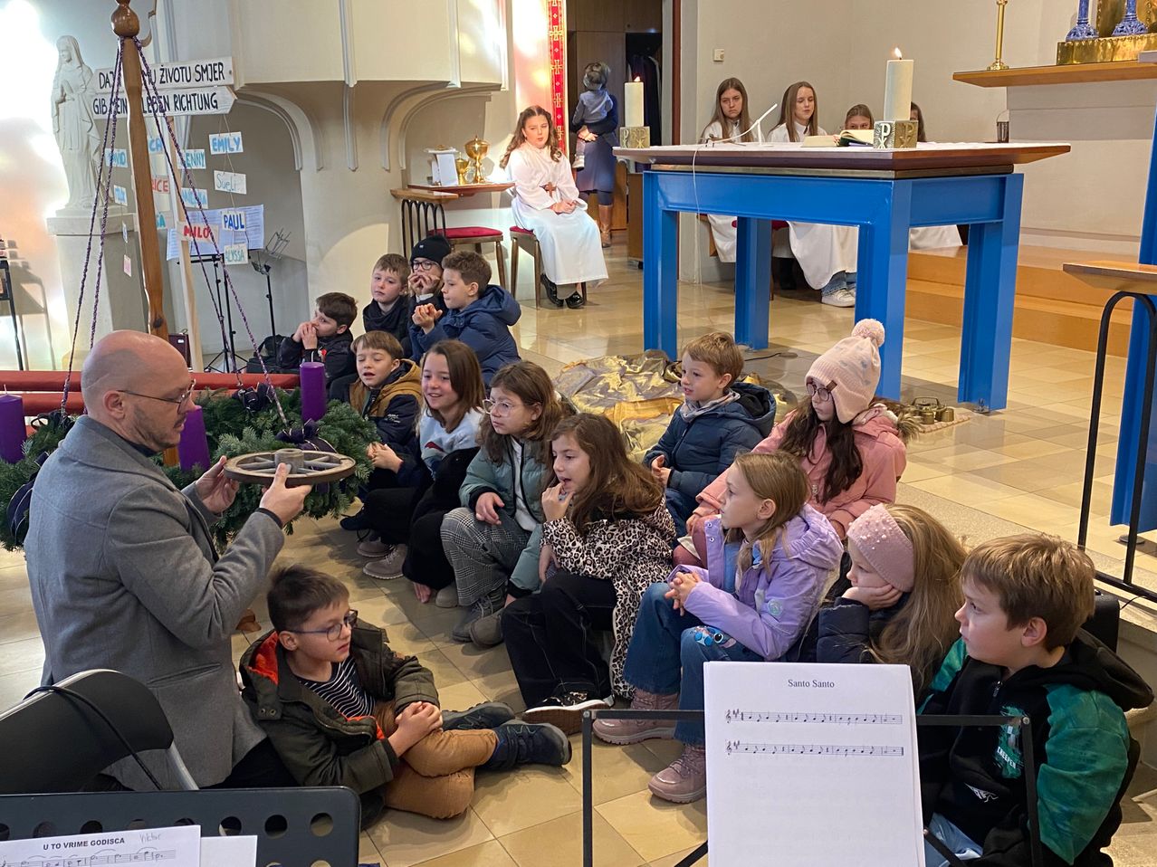 Eine Gruppe von Kindern und Erwachsenen sitzt um einen Tisch in einer Kirche herum. Eine Frau in Weiß steht neben dem Tisch, auf dem zwei Kerzen brennen. Die Kinder lauschen aufmerksam einem Mann, der ein Musikinstrument in der Hand hält.