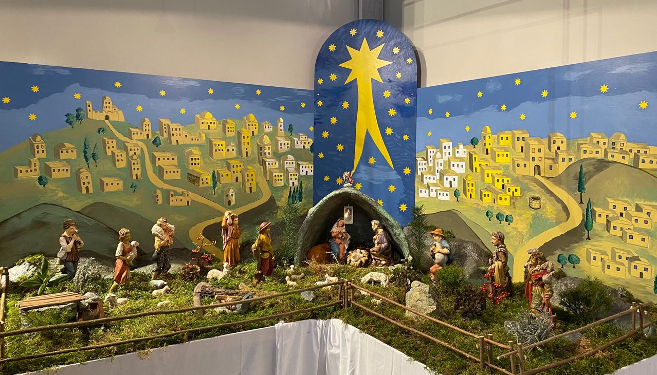 Eine lebendige Krippenszene mit einem Stern, Jesus, Maria und Josef in einer Höhle. Umgebende Figuren sind Hirten, Tiere und gemalte Bethlehem-Hintergründe.