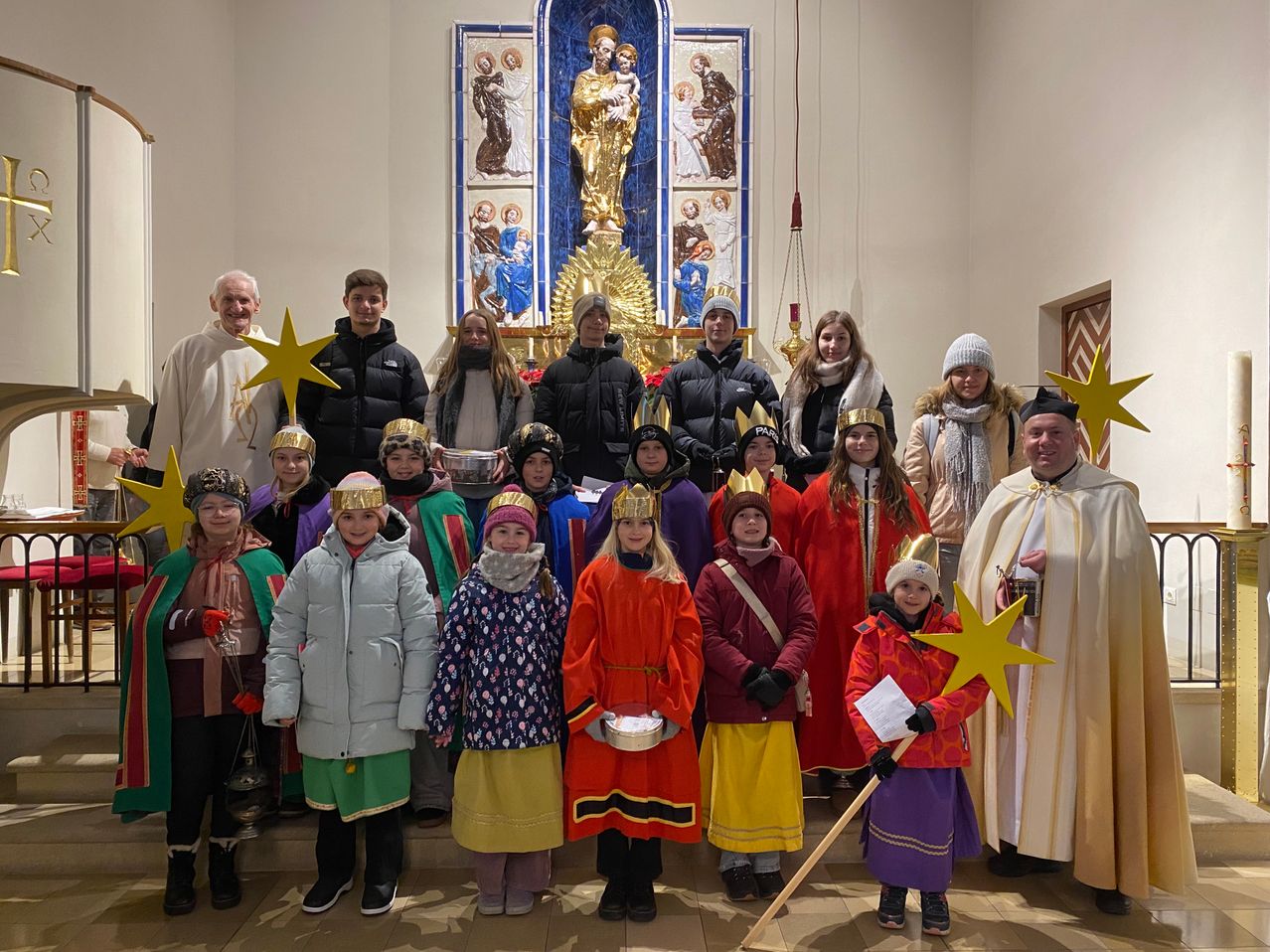 Eine Gruppe von Kindern und Erwachsenen in farbenfrohen Kostümen, einschließlich Kronen, steht vor einem Kirchenaltar. Sie halten Papiersterne und Schalen, und zwei Personen tragen weiße Gewänder. Hinter ihnen steht eine goldene Statue.