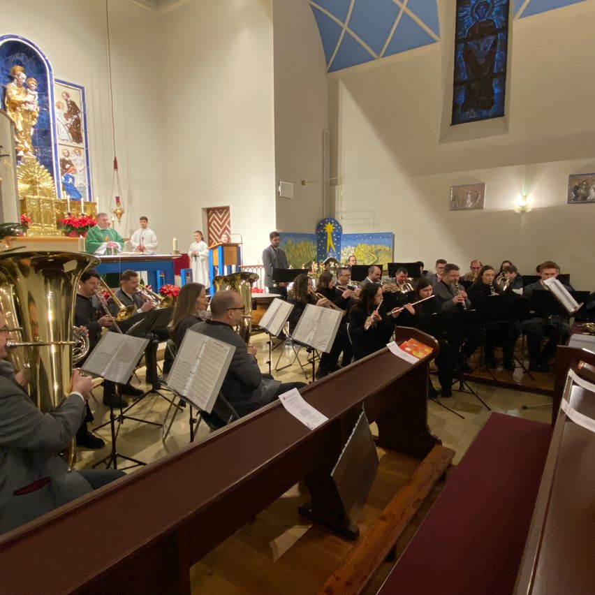 Eine Gruppe von Musikern spielt in einer Kirche, wobei verschiedene Instrumente sichtbar sind, darunter Tuba, Trompeten und Flöten. Die Musiker sitzen in Reihen, mit Notenständern vor ihnen. Dahinter steht ein Priester in der Nähe einer Kanzel.