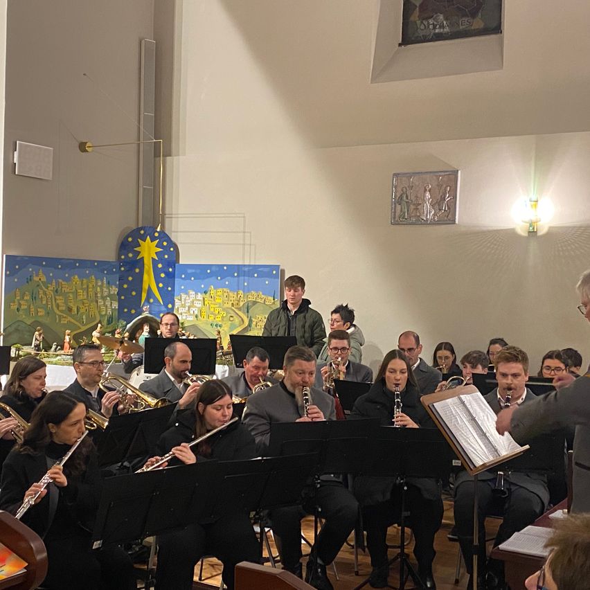Ein Orchester spielt in einer Kirche. Sie haben verschiedene Instrumente wie Trompeten, Flöten und Klarinetten. Die Orchestermitglieder sitzen auf Stühlen und halten ihre Instrumente. Der Dirigent steht vor dem Orchester. An der Wand hängen Gemälde, und ein Stern hängt von der Decke.