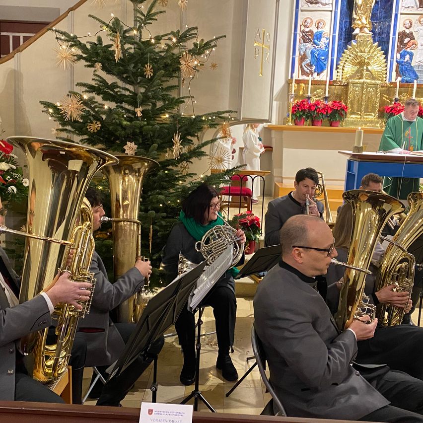 Ein Blechbläserensemble spielt in einer Kirche mit einem geschmückten Weihnachtsbaum. Die Musiker sitzen vor dem Altar, mit einem Priester hinter ihnen. Die Kirche ist mit religiösen Gemälden und Kerzen geschmückt.