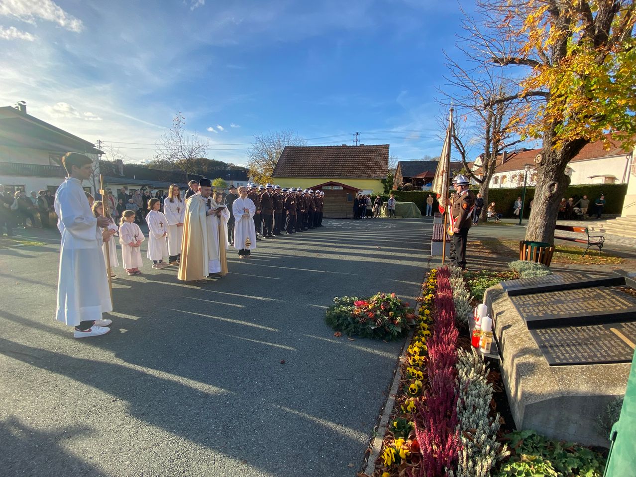 Eine feierliche Prozession von Geistlichen und Militärangehörigen steht auf einer Straße, in der Nähe eines Denkmals, unter blauem Himmel.