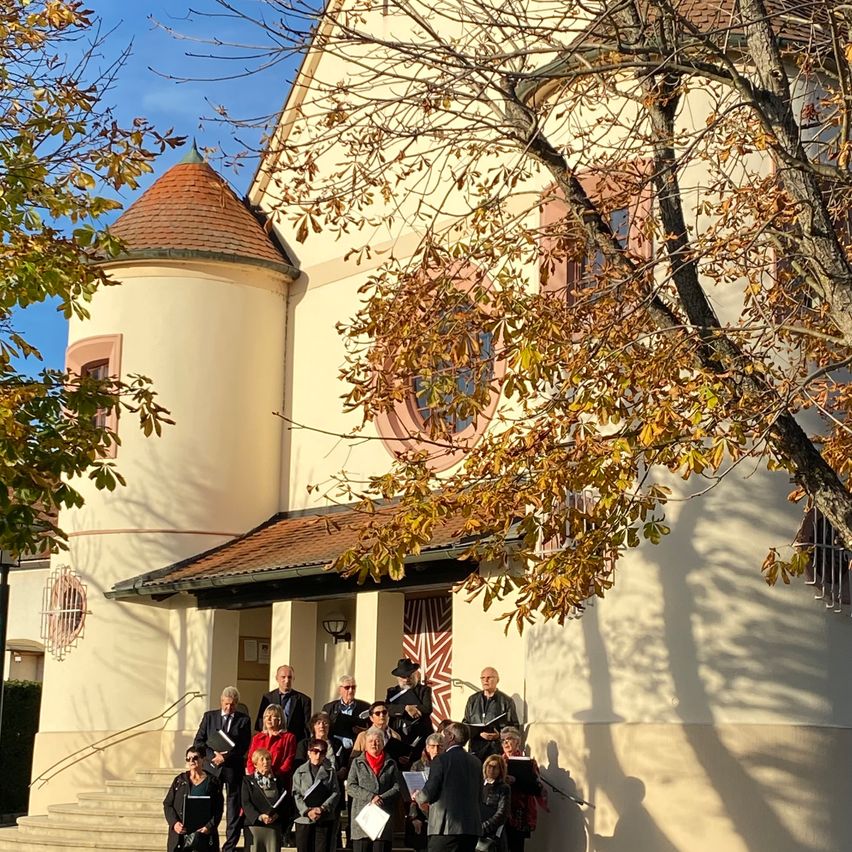 Eine Gruppe Erwachsener steht vor einer Kirche mit Herbstblättern an den Bäumen. Sie sind möglicherweise Chormitglieder, die auf eine Veranstaltung warten.