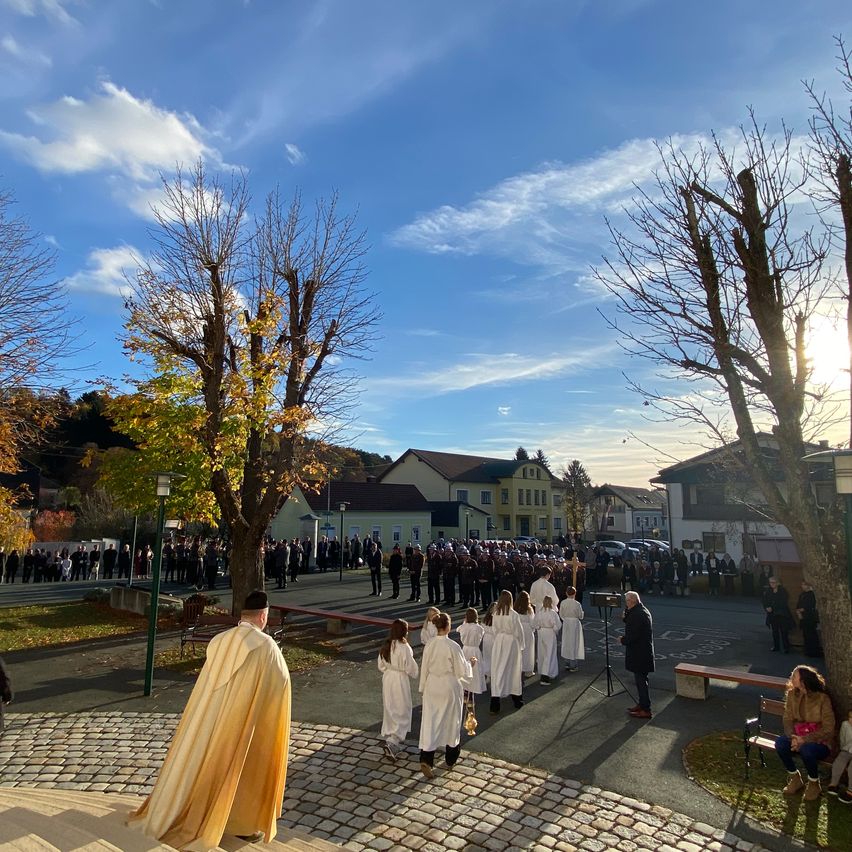 Eine große Gruppe versammelt sich im Freien für eine religiöse Zeremonie, die von einem Priester geleitet wird, viele Menschen in weißen Roben und einem klaren blauen Himmel im Hintergrund.